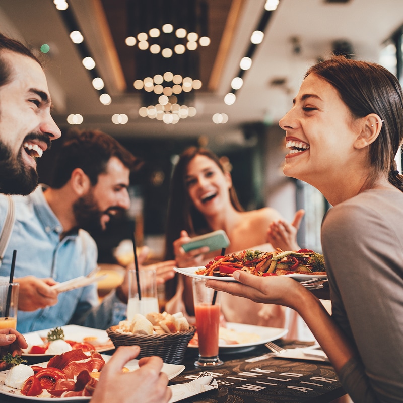 Groupe d'amis riant joyeusement autour d'une table de restaurant, partageant un repas avec salades, charcuteries et jus. Ambiance conviviale.