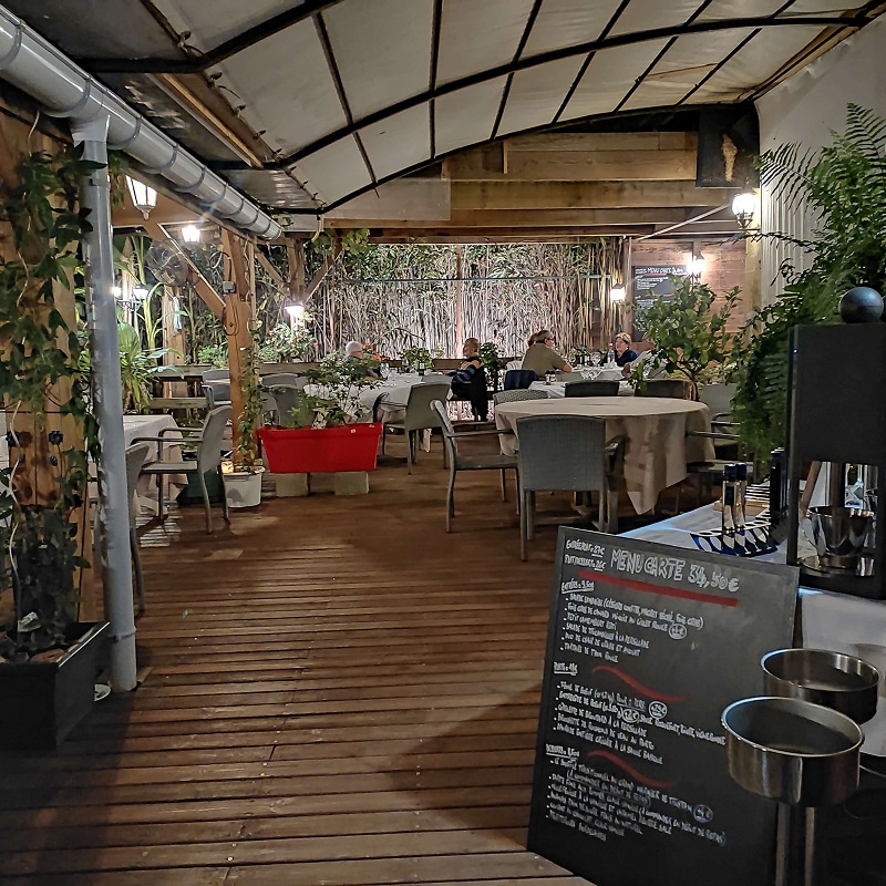 Restaurant : Terrasse chaleureuse en soirée, menu 34,50€ Terrasse de restaurant conviviale la nuit, sol en bois, tables dressées, plantes luxuriantes. Des clients dînent, ambiance chaleureuse. Menu affiché : 34,50€.